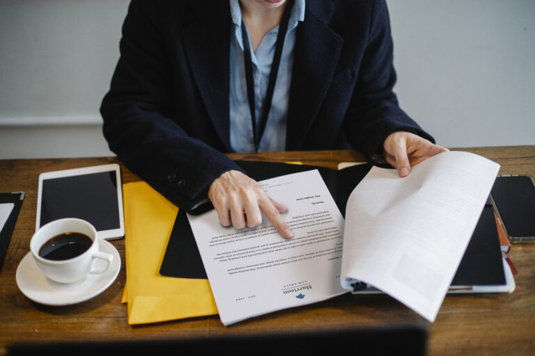 Personne en veste sombre qui parcourt des documents imprimés sur un bureau, à côté d'une tablette et d'une tasse de café